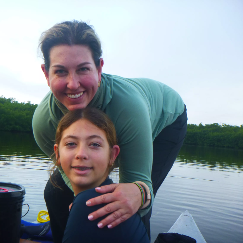 A woman and a girl are posing for a photo in what appears to be a boat or canoe. The woman is standing behind the girl with her arm around her. They are both smiling at the camera. The background shows a body of water and some trees, suggesting they might be on a lake or river. The sky is overcast, creating soft lighting.